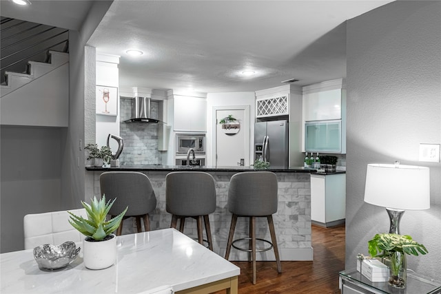 kitchen with white cabinetry, kitchen peninsula, wall chimney range hood, appliances with stainless steel finishes, and dark hardwood / wood-style flooring