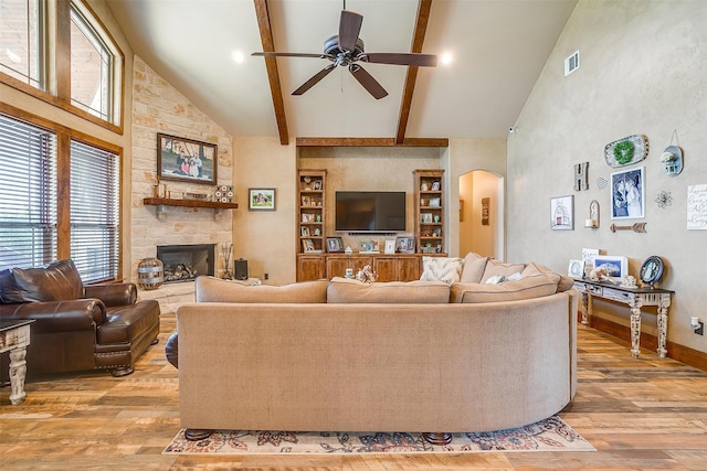 living room with ceiling fan, beam ceiling, a stone fireplace, high vaulted ceiling, and light wood-type flooring