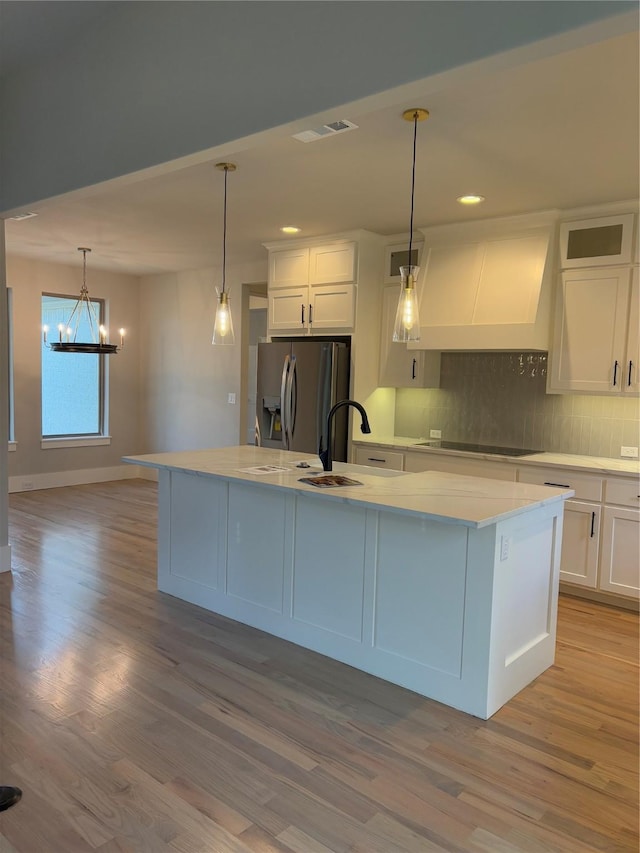 kitchen featuring white cabinetry, stainless steel refrigerator with ice dispenser, a center island with sink, black electric cooktop, and custom exhaust hood
