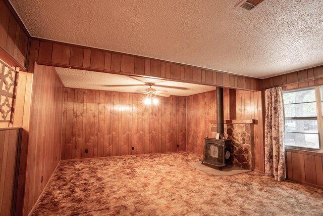 interior space with a textured ceiling, carpet, a wood stove, ceiling fan, and wood walls