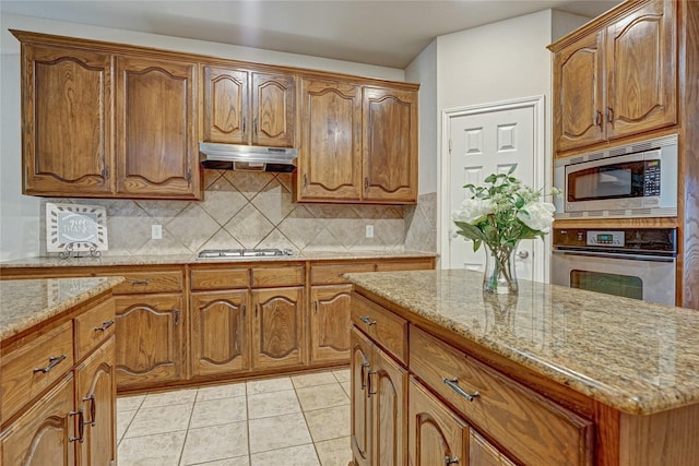 kitchen with light tile patterned floors, brown cabinetry, stainless steel appliances, under cabinet range hood, and backsplash