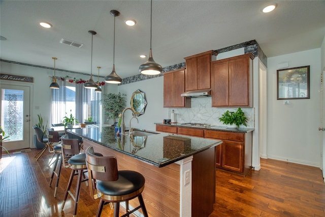 kitchen featuring decorative backsplash, sink, a center island with sink, decorative light fixtures, and dark hardwood / wood-style flooring