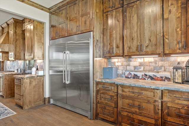 kitchen with tasteful backsplash, built in refrigerator, light stone counters, dark wood-style floors, and brown cabinets