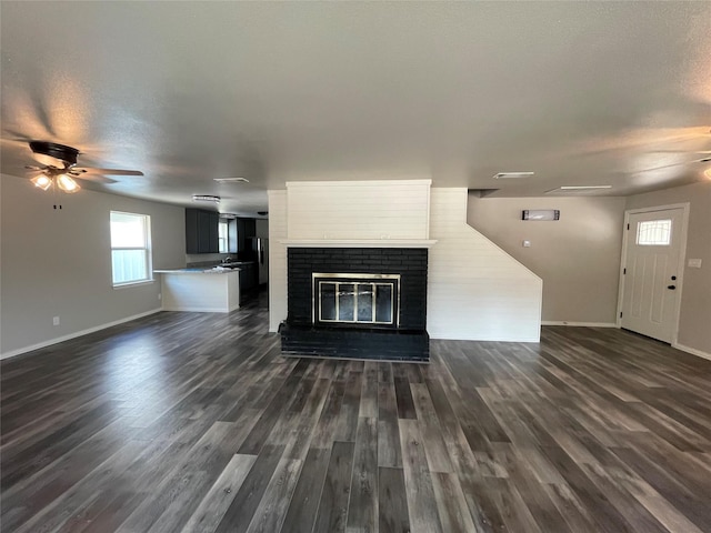 unfurnished living room featuring dark wood-style floors, a brick fireplace, ceiling fan, a textured ceiling, and baseboards