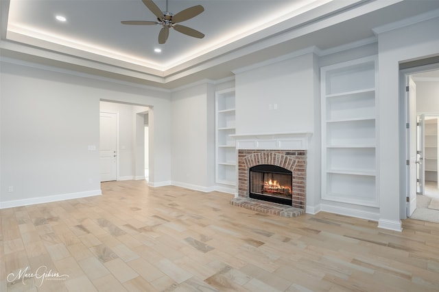 unfurnished living room with a brick fireplace, light wood-type flooring, built in features, ceiling fan, and a raised ceiling