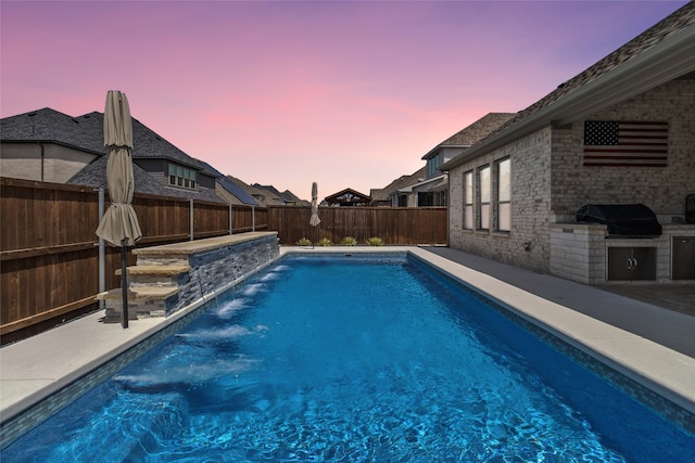 pool at dusk with grilling area, an outdoor kitchen, and pool water feature