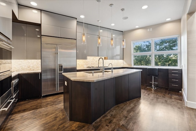 kitchen with dark wood-type flooring, stainless steel built in refrigerator, sink, tasteful backsplash, and decorative light fixtures