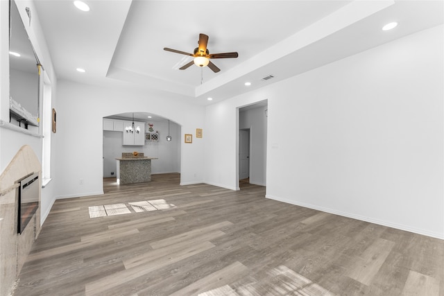 unfurnished living room featuring wood-type flooring, a tray ceiling, a tiled fireplace, and ceiling fan with notable chandelier