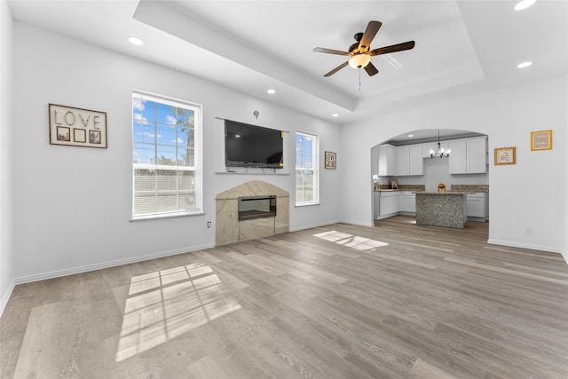 unfurnished living room featuring a raised ceiling, a premium fireplace, ceiling fan with notable chandelier, and light wood-type flooring