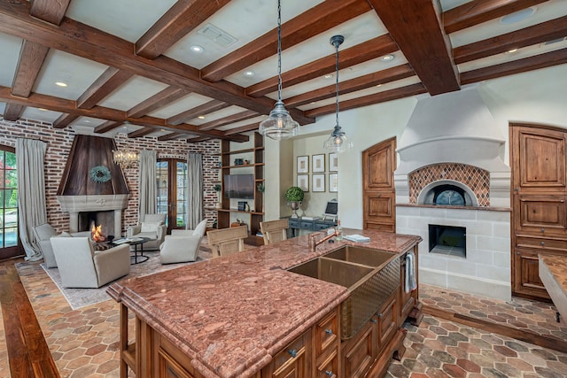 kitchen featuring decorative light fixtures, beamed ceiling, an island with sink, a large fireplace, and sink