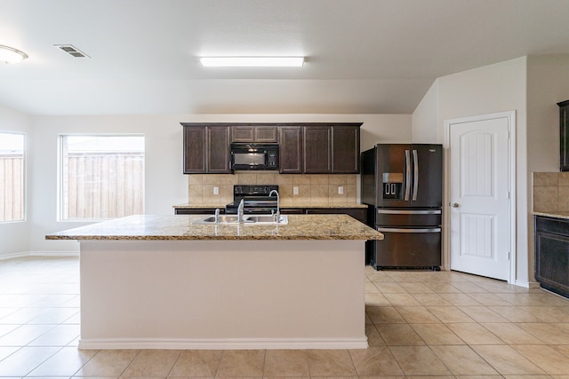 kitchen with light tile patterned floors, an island with sink, backsplash, black appliances, and light stone counters