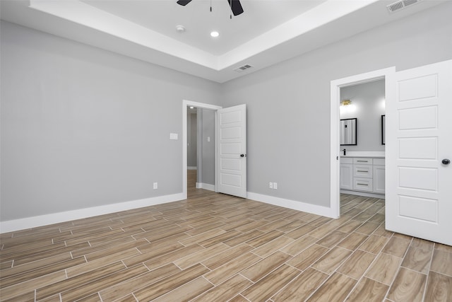 unfurnished bedroom with ceiling fan, ensuite bath, a raised ceiling, and light hardwood / wood-style floors