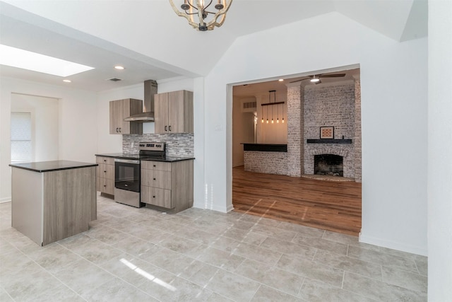 kitchen featuring lofted ceiling with skylight, a fireplace, light brown cabinetry, stainless steel electric stove, and wall chimney exhaust hood