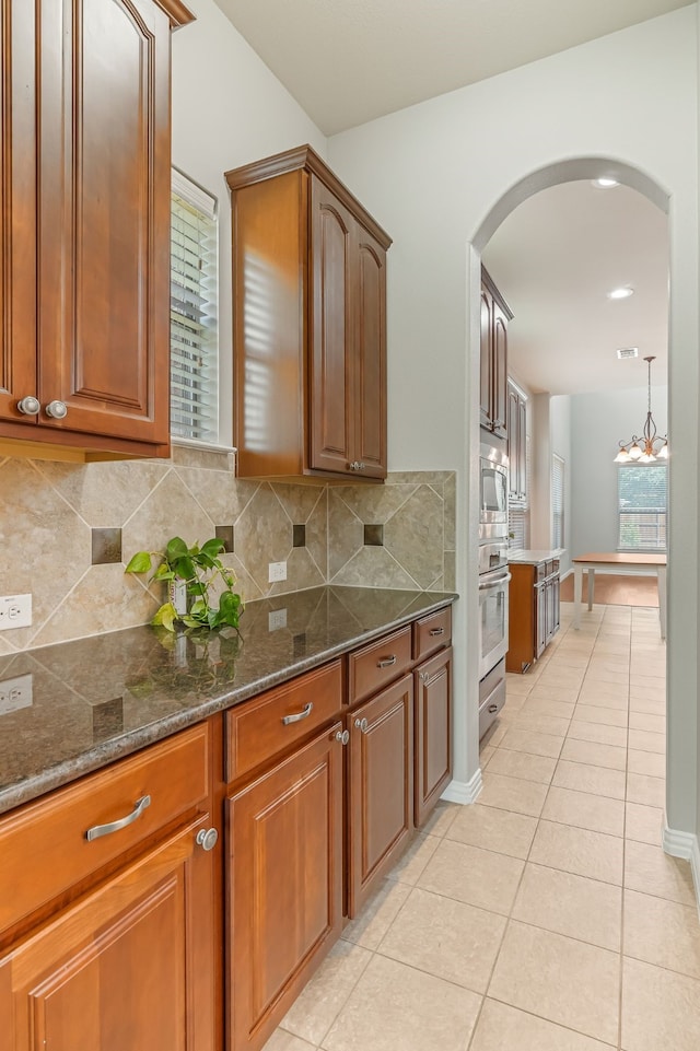 kitchen with tasteful backsplash, brown cabinetry, light tile patterned floors, arched walkways, and stainless steel appliances