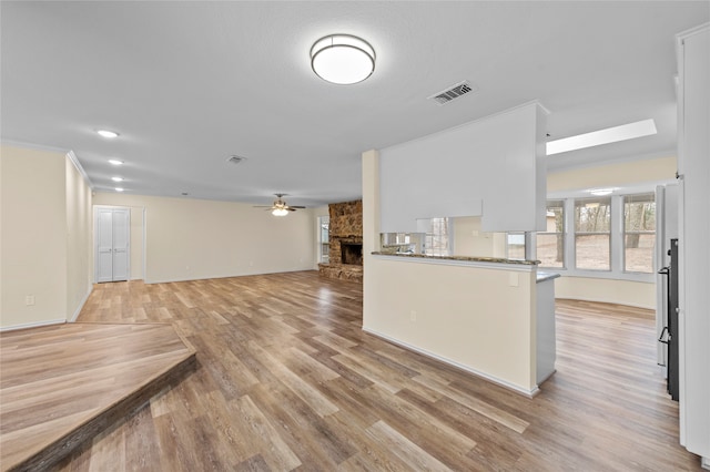 unfurnished living room featuring a skylight, light wood-type flooring, ceiling fan, crown molding, and a stone fireplace