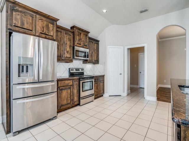 kitchen with dark stone countertops, arched walkways, appliances with stainless steel finishes, light tile patterned floors, and decorative backsplash