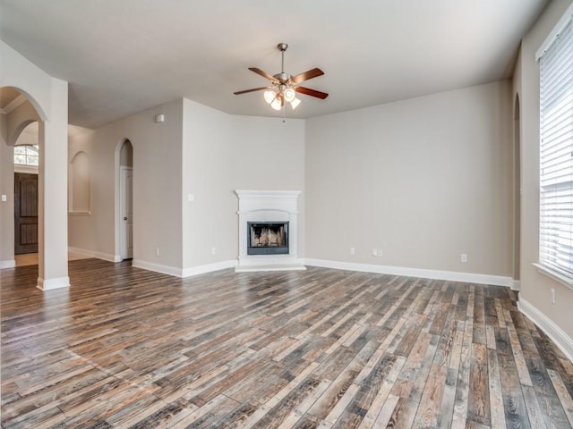 unfurnished living room with a fireplace with raised hearth, a ceiling fan, wood finished floors, arched walkways, and baseboards