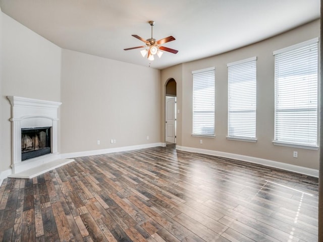 unfurnished living room with a fireplace with raised hearth, baseboards, ceiling fan, wood finished floors, and arched walkways