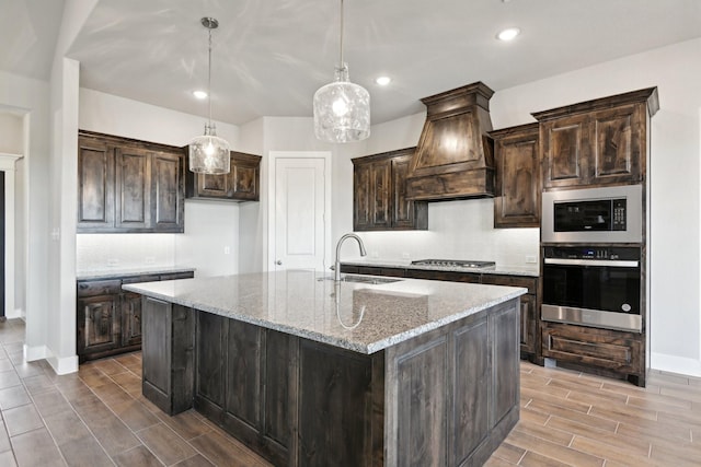 kitchen with a center island with sink, stainless steel appliances, hanging light fixtures, light stone counters, and sink