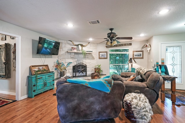 living room with a textured ceiling, light wood-type flooring, a stone fireplace, and ceiling fan