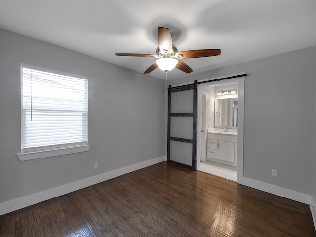 unfurnished bedroom with ceiling fan, connected bathroom, a barn door, and dark hardwood / wood-style flooring