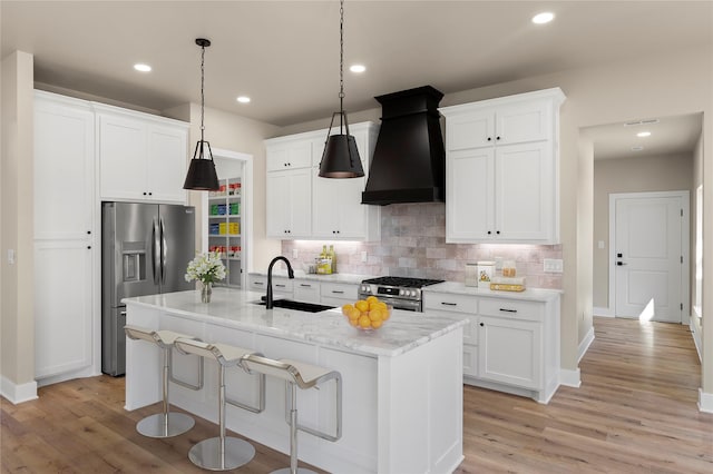 kitchen with sink, white cabinetry, appliances with stainless steel finishes, custom range hood, and a kitchen island with sink