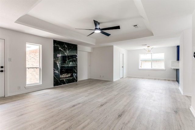 unfurnished living room featuring a raised ceiling, a high end fireplace, ceiling fan with notable chandelier, and light hardwood / wood-style floors