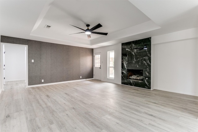 unfurnished living room featuring a raised ceiling, ceiling fan, a premium fireplace, and light hardwood / wood-style floors