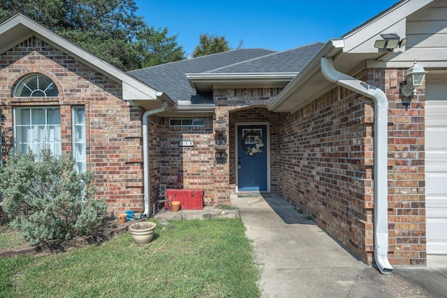 doorway to property featuring an attached garage, a yard, roof with shingles, and brick siding