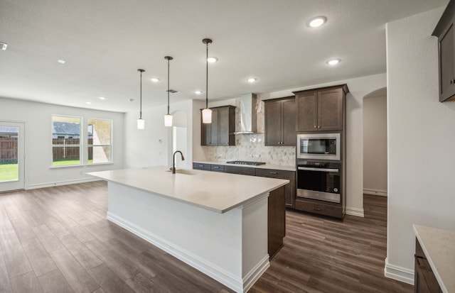 kitchen featuring wall chimney range hood, hanging light fixtures, a center island with sink, dark hardwood / wood-style flooring, and stainless steel appliances