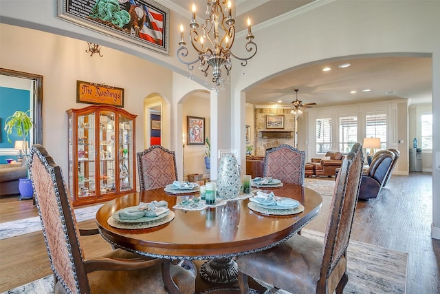 dining space featuring arched walkways, a stone fireplace, ceiling fan with notable chandelier, wood finished floors, and crown molding