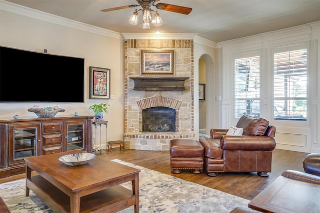 living area featuring a ceiling fan, ornamental molding, wood finished floors, and a stone fireplace