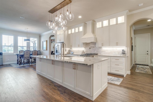 kitchen with arched walkways, a kitchen island with sink, premium range hood, white cabinets, and glass insert cabinets