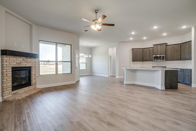 kitchen with ceiling fan with notable chandelier, light hardwood / wood-style flooring, a fireplace, an island with sink, and dark brown cabinetry