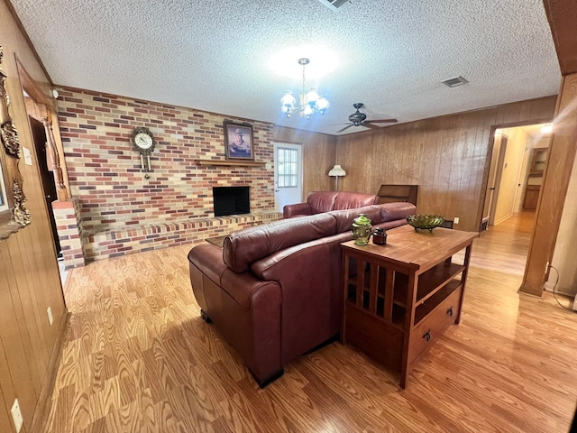 living room featuring light hardwood / wood-style floors, ceiling fan with notable chandelier, wood walls, a fireplace, and brick wall