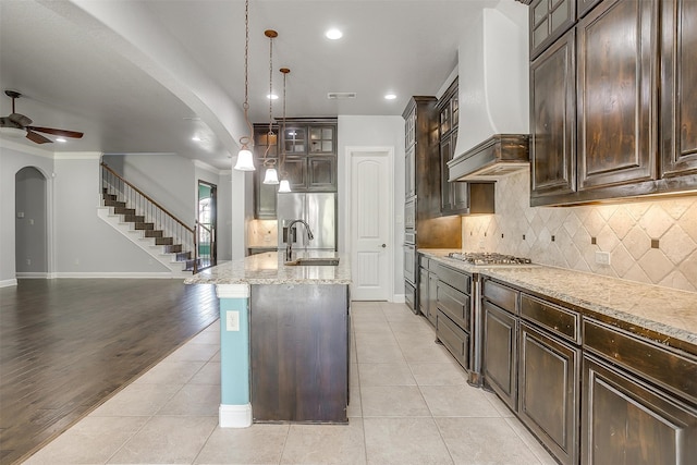 kitchen with ceiling fan, pendant lighting, custom range hood, a center island with sink, and light hardwood / wood-style floors