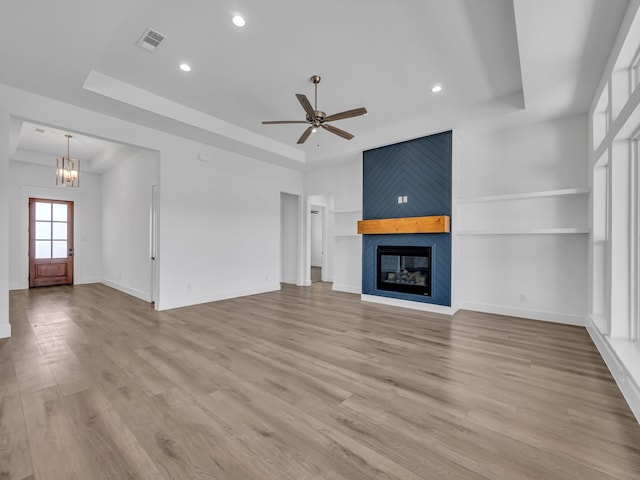 unfurnished living room with built in shelves, light wood-type flooring, a large fireplace, a tray ceiling, and ceiling fan with notable chandelier