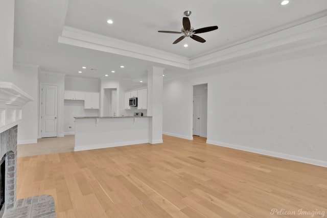 unfurnished living room with crown molding, a fireplace, ceiling fan, a tray ceiling, and light hardwood / wood-style flooring