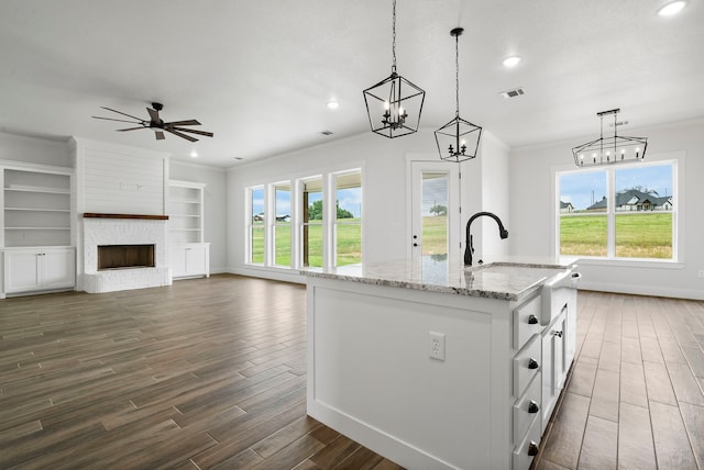 kitchen featuring a kitchen island with sink, a brick fireplace, decorative light fixtures, white cabinets, and light stone counters