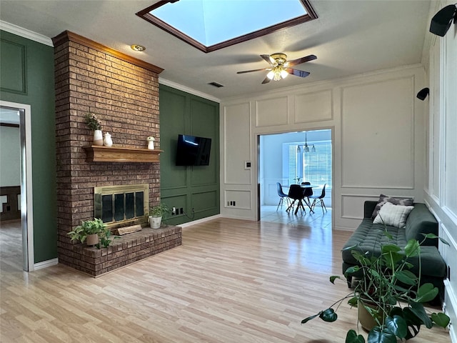 living room featuring light hardwood / wood-style flooring, ornamental molding, a skylight, ceiling fan, and a brick fireplace