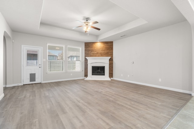 unfurnished living room with a large fireplace, a tray ceiling, light wood-type flooring, and ceiling fan