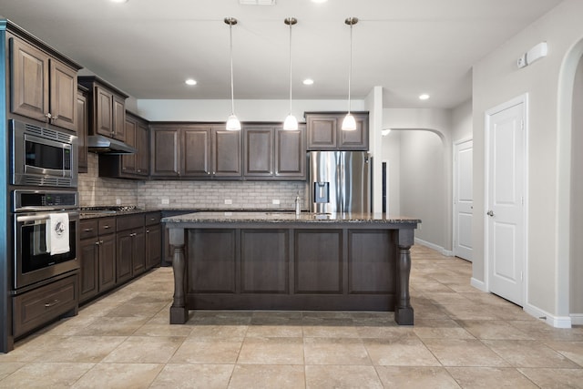 kitchen with appliances with stainless steel finishes, an island with sink, dark brown cabinets, dark stone counters, and decorative light fixtures