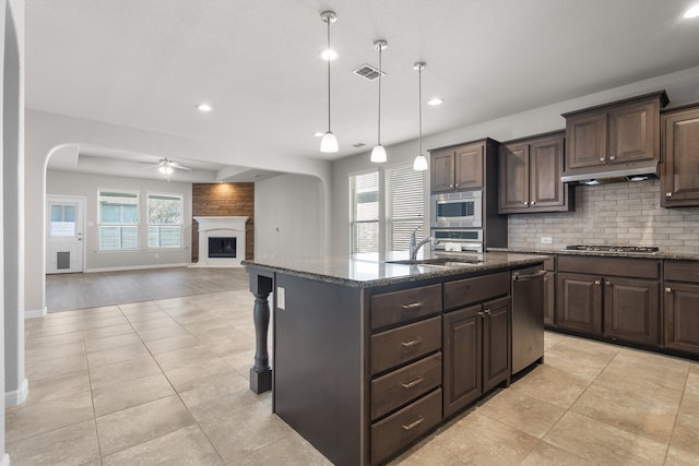 kitchen with a large fireplace, hanging light fixtures, a kitchen island with sink, dark brown cabinetry, and stainless steel appliances