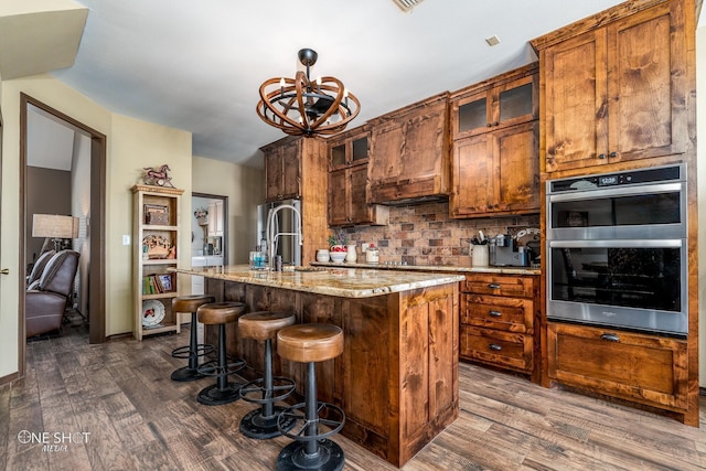 kitchen featuring double oven, dark hardwood / wood-style floors, an island with sink, light stone countertops, and tasteful backsplash