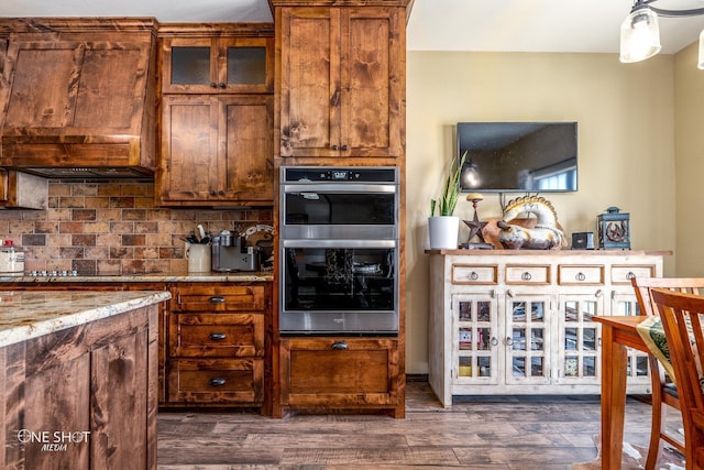 kitchen with stainless steel double oven, black electric cooktop, custom range hood, light stone counters, and dark hardwood / wood-style floors