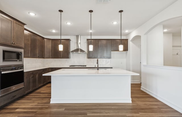 kitchen featuring a center island with sink, wall chimney range hood, sink, hanging light fixtures, and appliances with stainless steel finishes