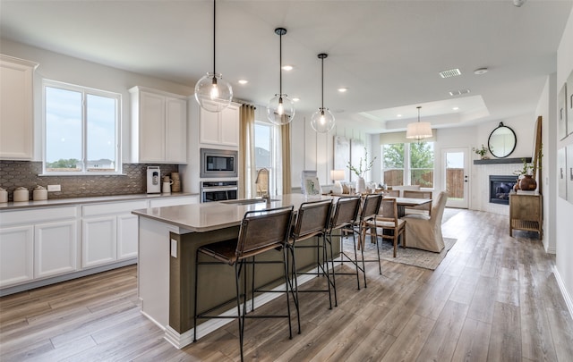 kitchen featuring stainless steel appliances, a raised ceiling, an island with sink, light wood-type flooring, and a tiled fireplace