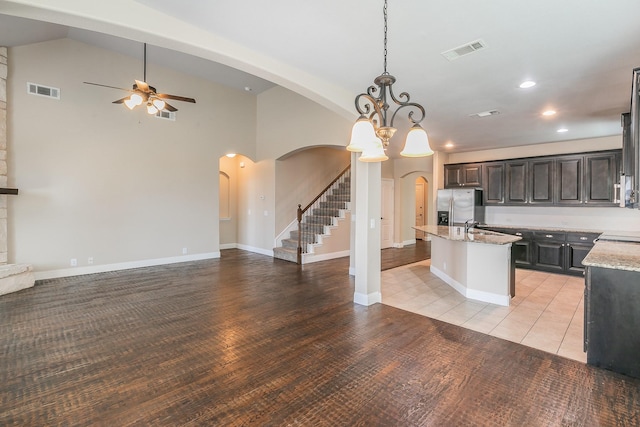 kitchen with stainless steel refrigerator with ice dispenser, dark brown cabinetry, a center island with sink, pendant lighting, and light stone countertops