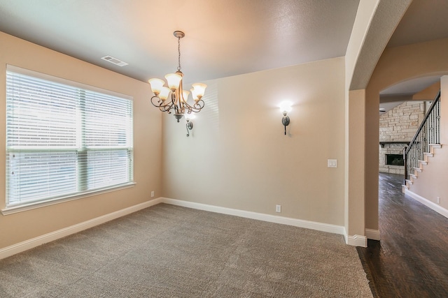 unfurnished room with dark colored carpet, a stone fireplace, and an inviting chandelier