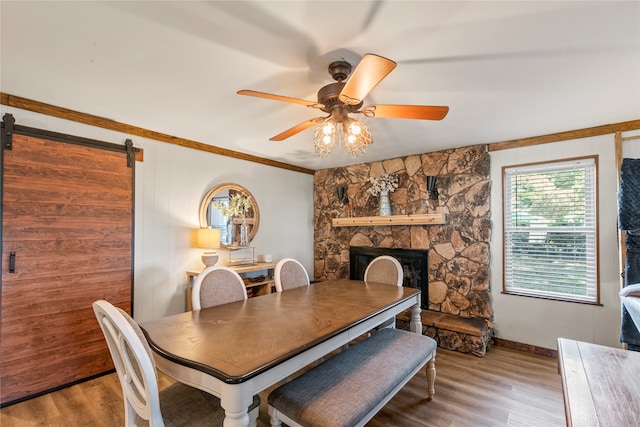 dining room with crown molding, wood-type flooring, a barn door, ceiling fan, and a stone fireplace